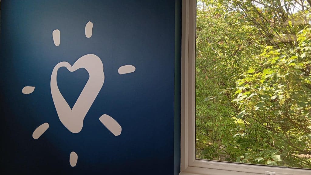 A white heart painted on a dark blue wall beside a window with greenery outside.