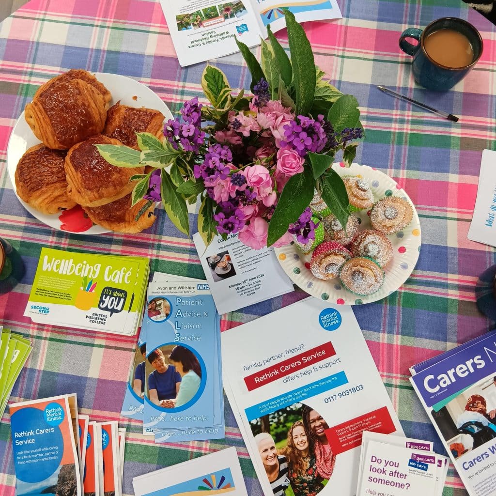 A flat lay of a table with a checked table cloth. On the table is a potted plant with pink flowers, a full mug of tea, two plates of pastries and piles of leaflets showing information about support for carers, including from Second Step's Bristol Wellbeing College, Rethink Mental Illness and Avon and Wiltshire Partnership