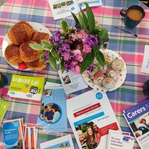 A flat lay of a table with a checked table cloth. On the table is a potted plant with pink flowers, a full mug of tea, two plates of pastries and piles of leaflets showing information about support for carers, including from Second Step's Bristol Wellbeing College, Rethink Mental Illness and Avon and Wiltshire Partnership