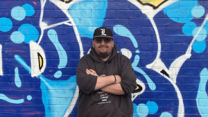 Drew standing arms crossed in front of a wall with colourful artwork on