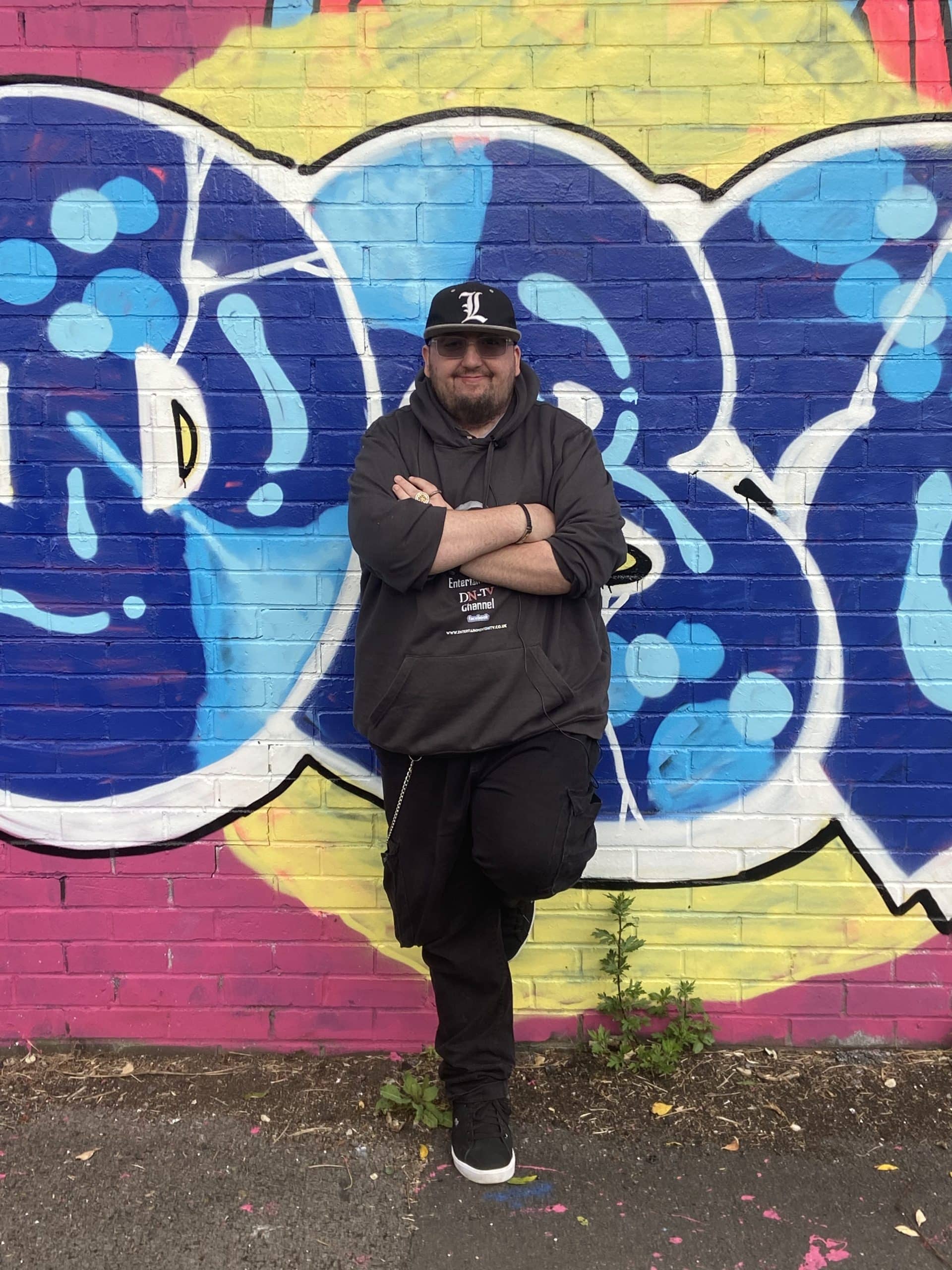 Drew standing arms crossed in front of a wall with colourful artwork on